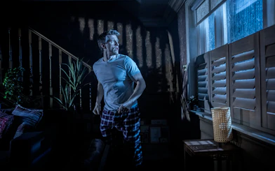 Man in a dimly lit room looking out a window during Paranormal Activity stage performance, Westend.