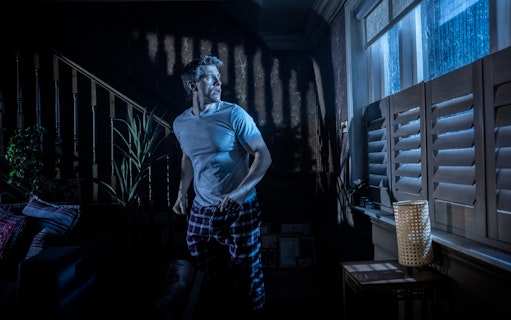 Man in a dimly lit room looking out a window during Paranormal Activity stage performance, Westend.