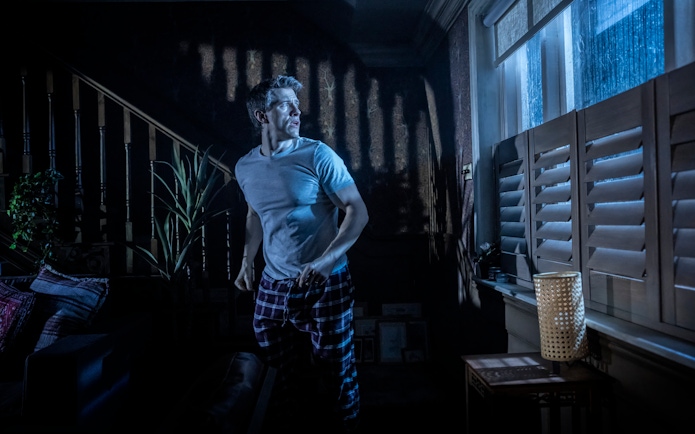 Man in a dimly lit room looking out a window during Paranormal Activity stage performance, Westend.
