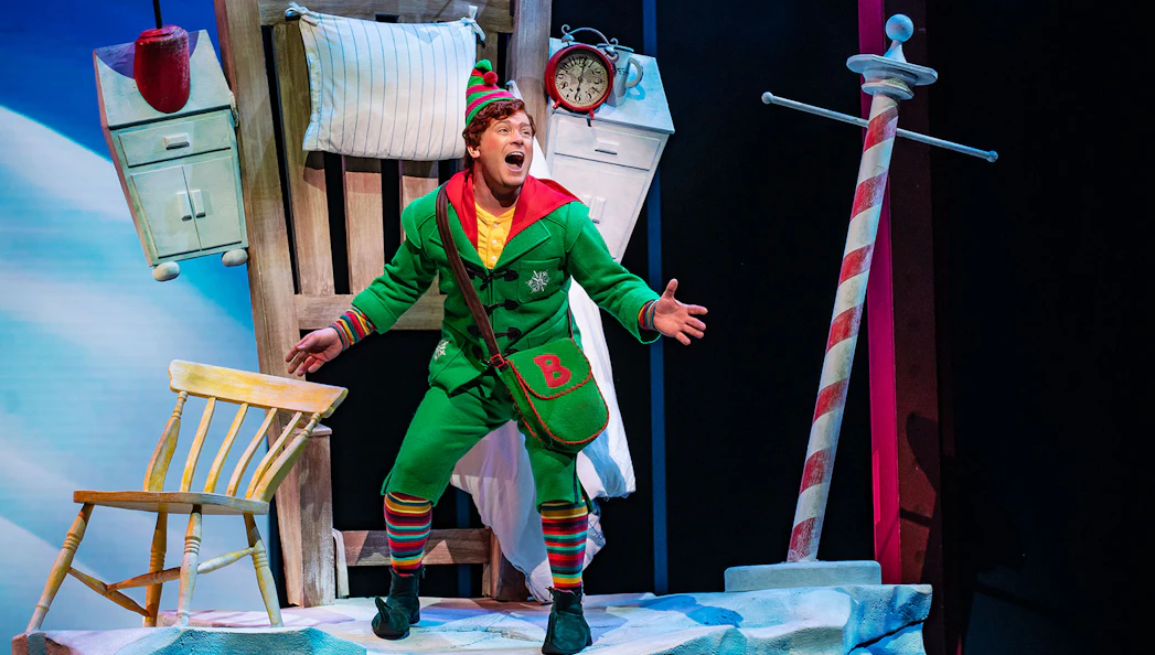 Elf character performing on stage in "Elf The Musical" with festive set design.