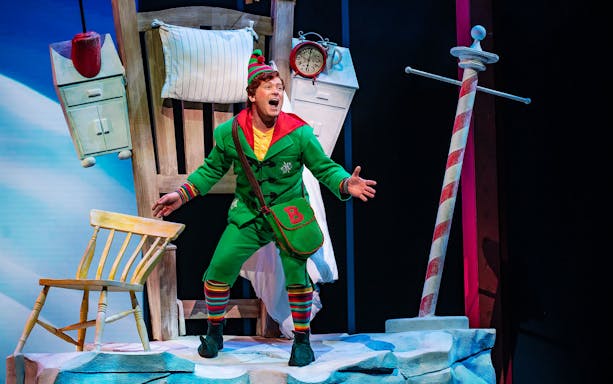 Elf character performing on stage in "Elf The Musical" with festive set design.