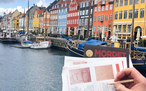 Nyhavn canal with colorful buildings and a person holding a mystery adventure booklet in Copenhagen.