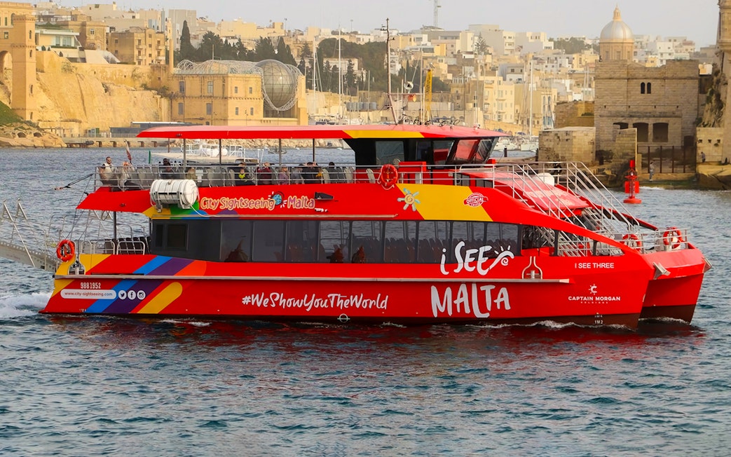 Red sightseeing boat cruising in Valletta harbor, Malta, with historic buildings in the background.