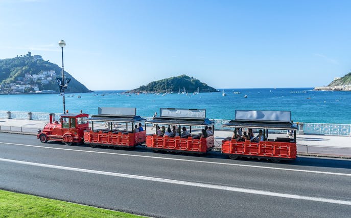 Red tourist train along San Sebastian coastline with ocean and hills in the background.