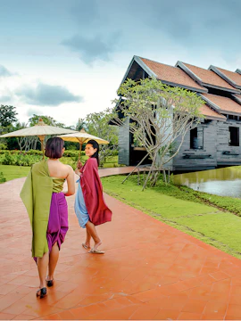 Women in traditional attire walking with umbrellas near a wooden building in Mallika City.