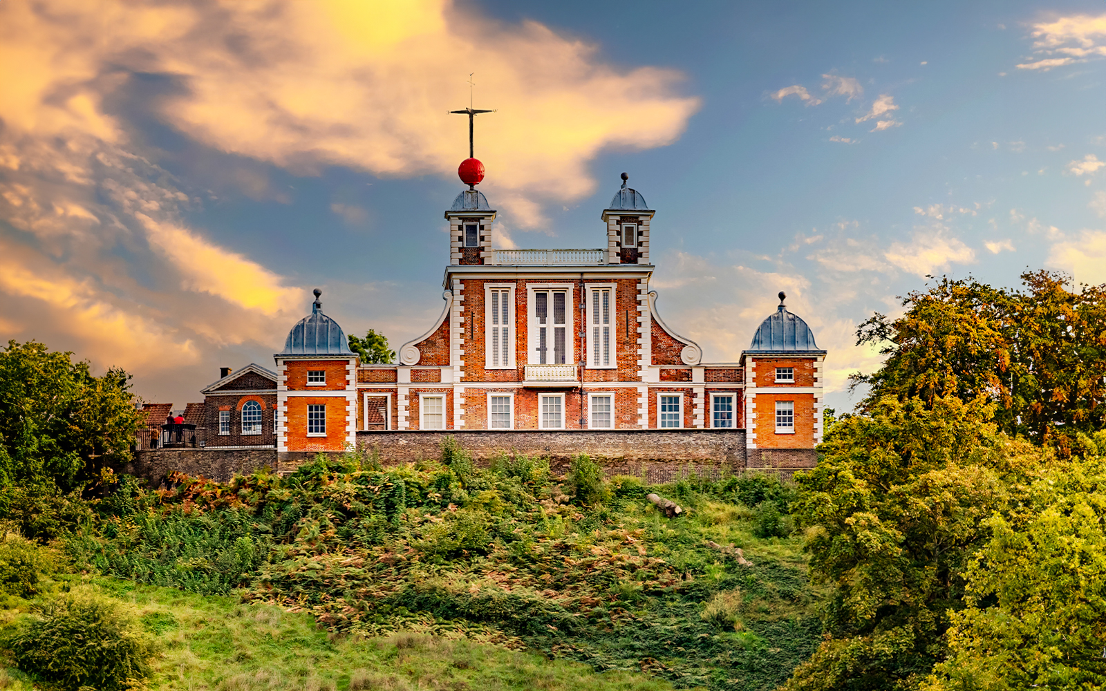 Royal Observatory Greenwich with red brick facade and domed towers against a sunset sky.