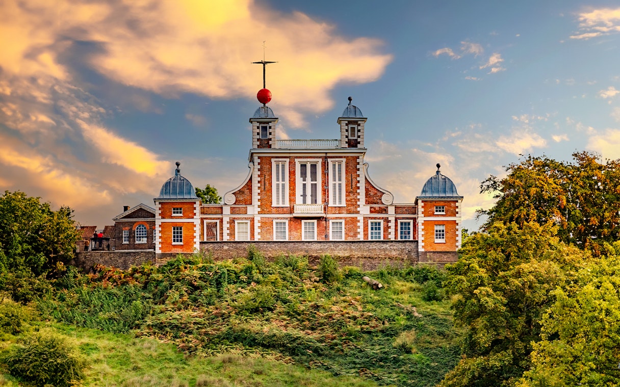 Royal Observatory Greenwich with red brick facade and domed towers against a sunset sky.