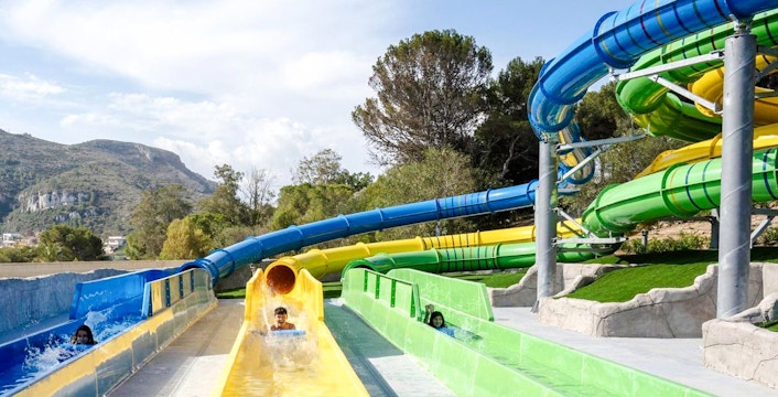 Guests enjoying water slides at Aquopolis Cullera with scenic mountain backdrop.