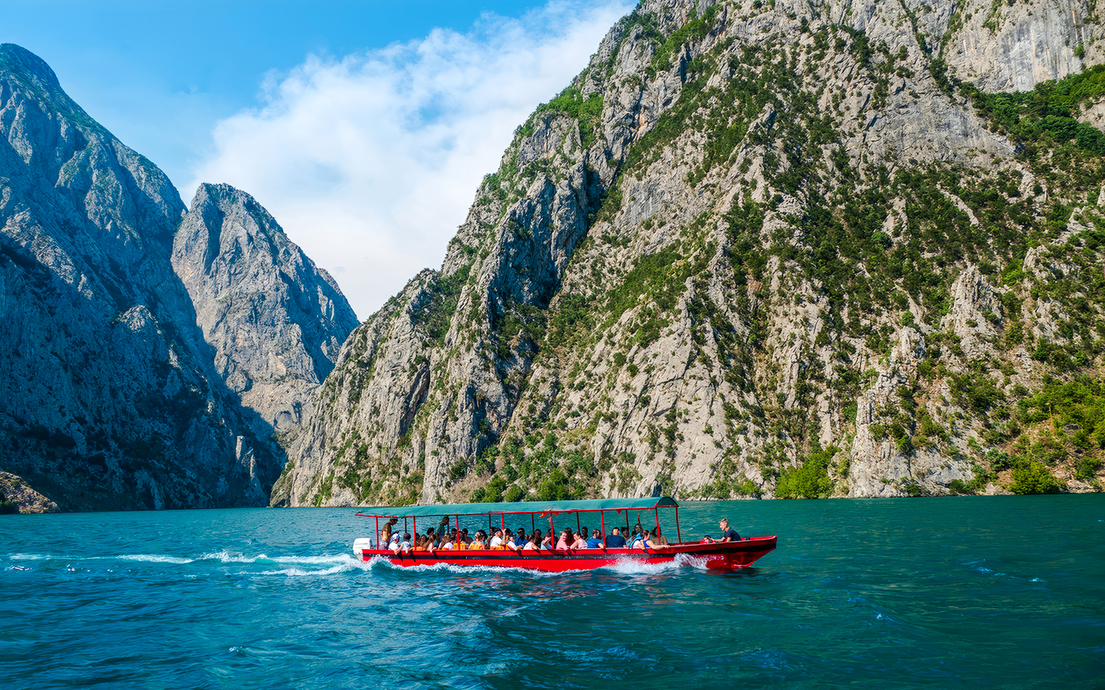 Speedboat on Koman Lake with passengers, surrounded by rocky cliffs and clear blue water.