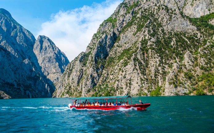 Speedboat on Koman Lake with passengers, surrounded by rocky cliffs and clear blue water.