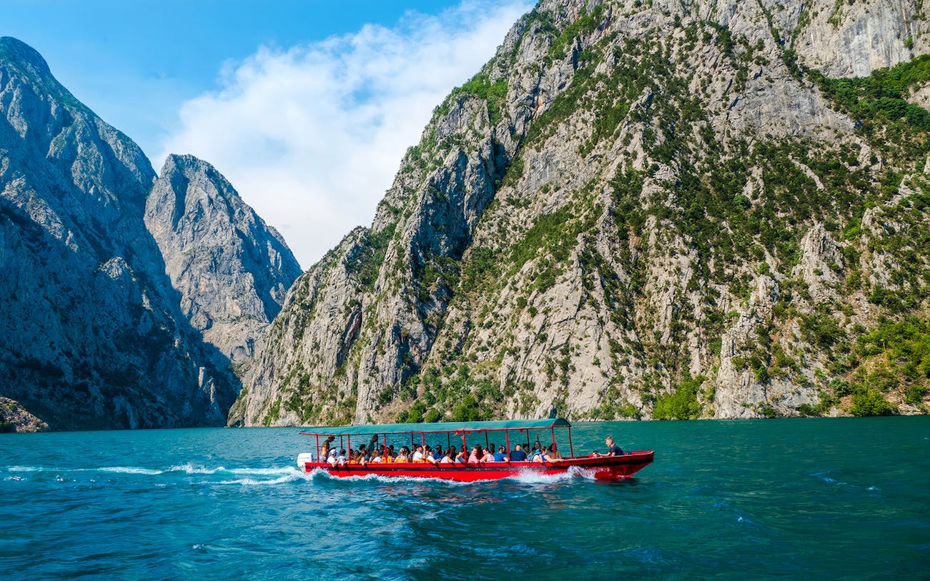 Speedboat on Koman Lake with passengers, surrounded by rocky cliffs and clear blue water.