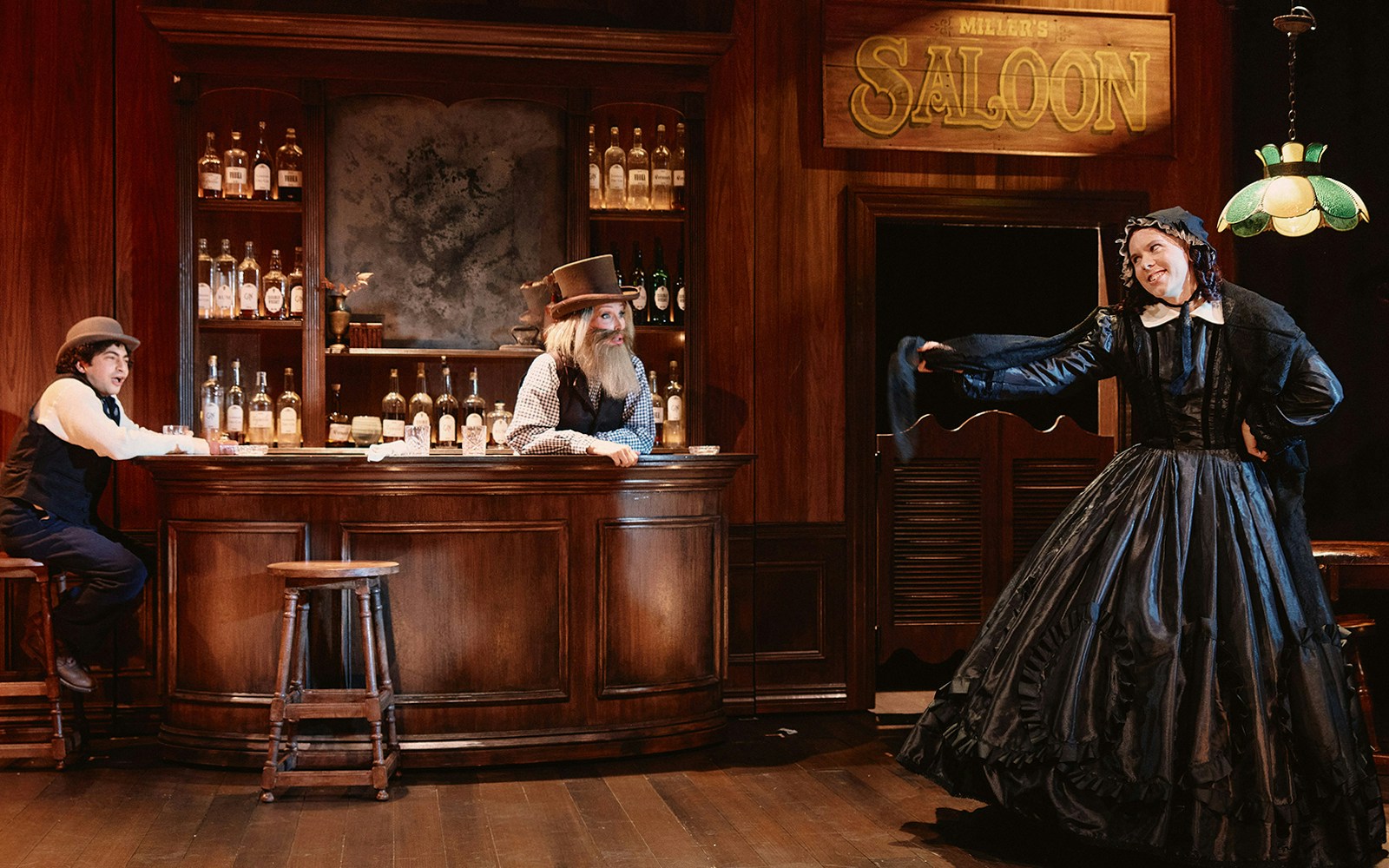 Saloon scene with actors in period costumes, featuring a woman in a black dress and two men at the bar.