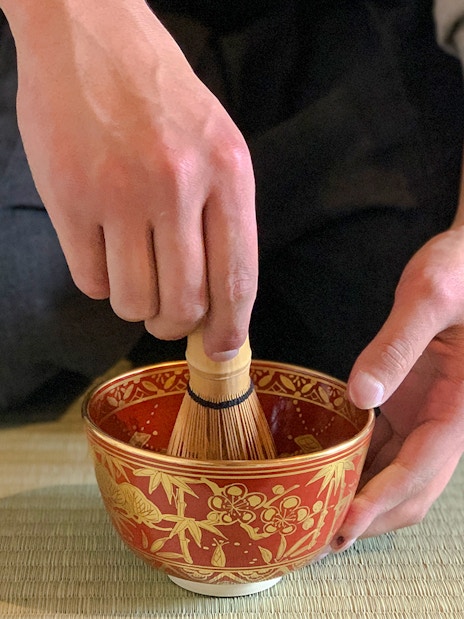 Whisking matcha green tea in a decorative bowl on a tatami mat.