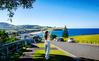 Person enjoying coastal view near Kiama Blowhole, with ocean and hills in the background, Sydney tour.