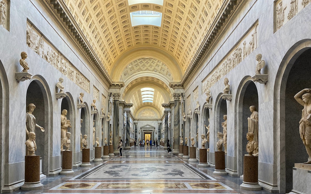 Tourists exploring the Vatican Museums' ornate hallway with classical sculptures in Rome, Italy.
