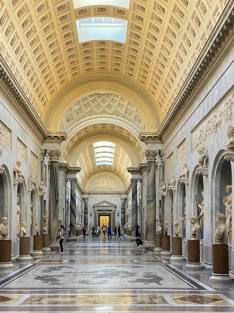 Tourists exploring the Vatican Museums' ornate hallway with classical sculptures in Rome, Italy.