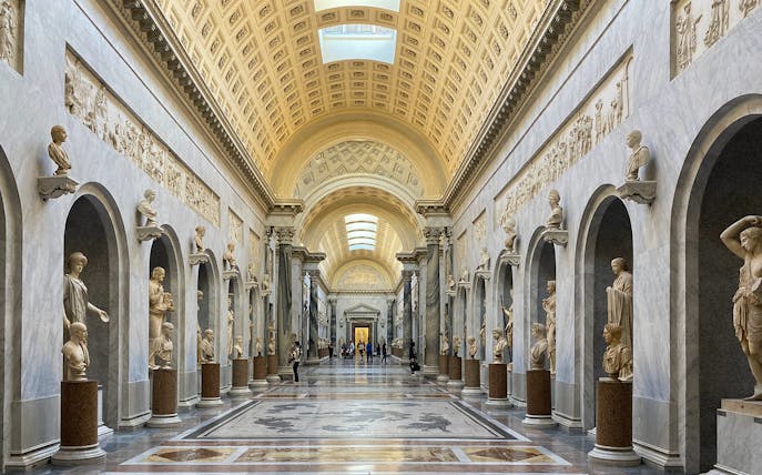 Tourists exploring the Vatican Museums' ornate hallway with classical sculptures in Rome, Italy.