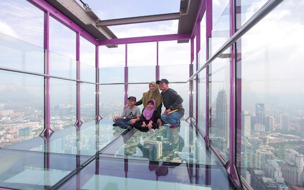 Family on glass sky deck at KL Tower with city view.