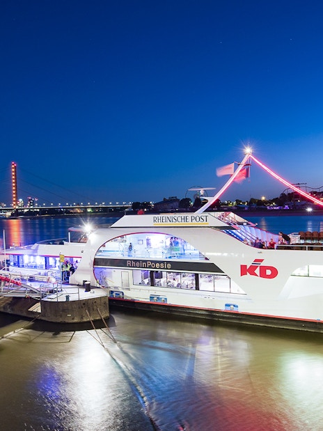 Cruise ship docked on the Rhine River in Düsseldorf with city skyline and Rheinturm Tower.