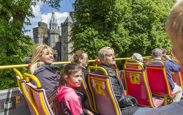 Open-top bus with tourists viewing Cardiff Castle on a sunny day.