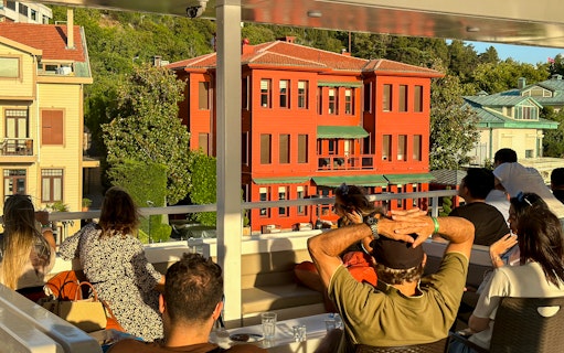 Tourists on a Bosphorus cruise viewing historic waterfront mansions during sunset.