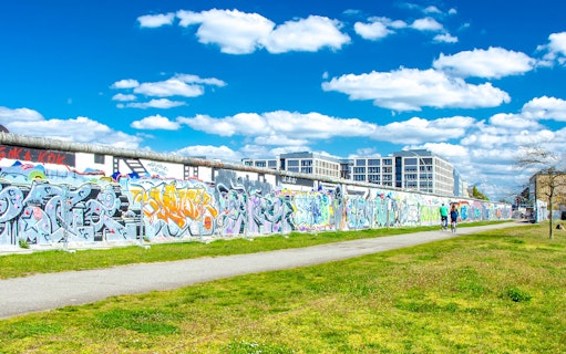 Cyclists ride past graffiti-covered Berlin Wall on a sunny day.