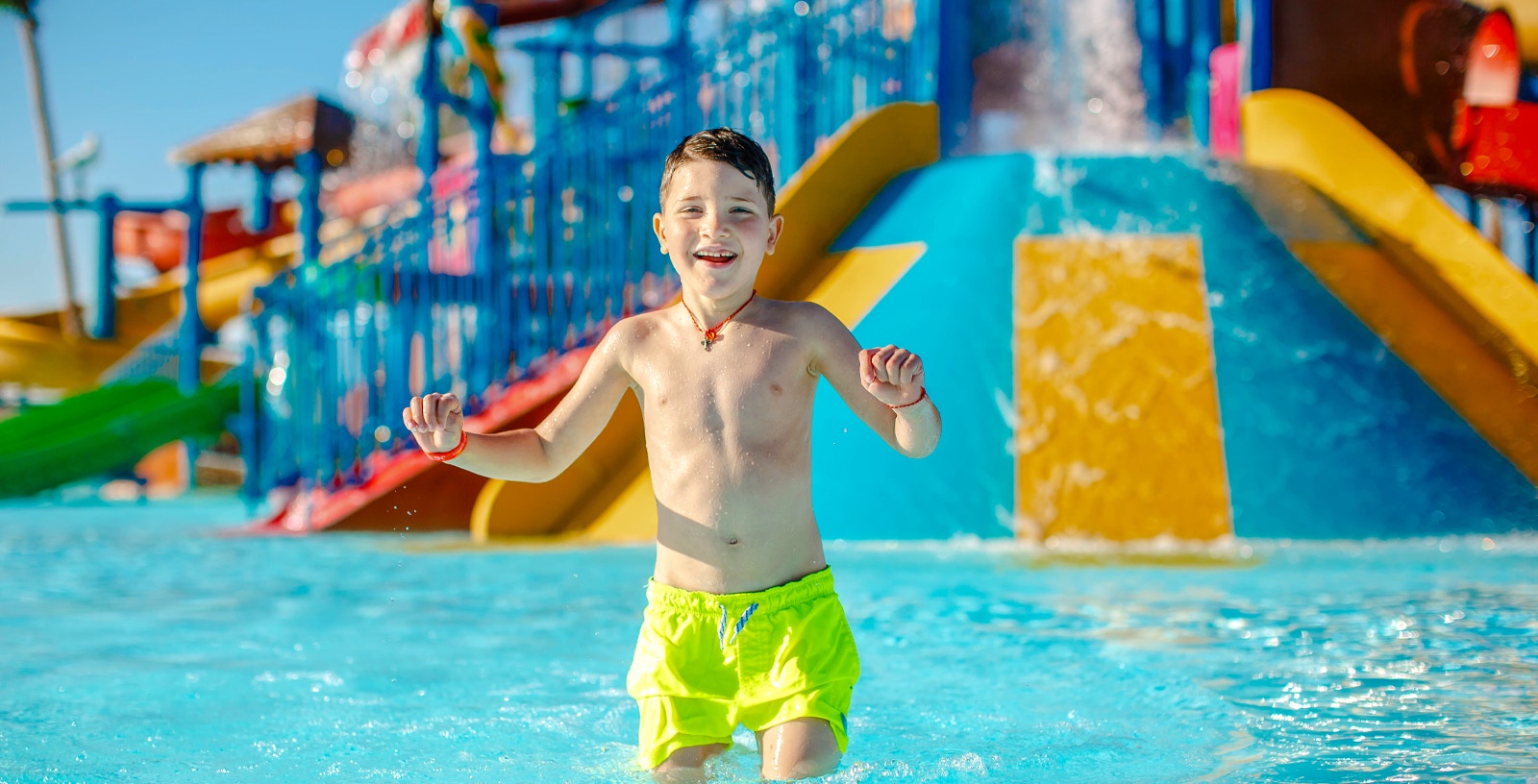 Child playing in a water park pool with colorful slides in the background.