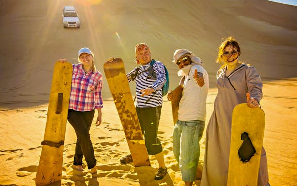 Group enjoying sandboarding during evening desert safari with BBQ dinner.