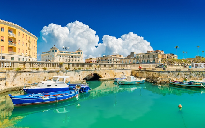 Boats docked in the turquoise waters of Siracusa, Italy, with historic buildings in the background.