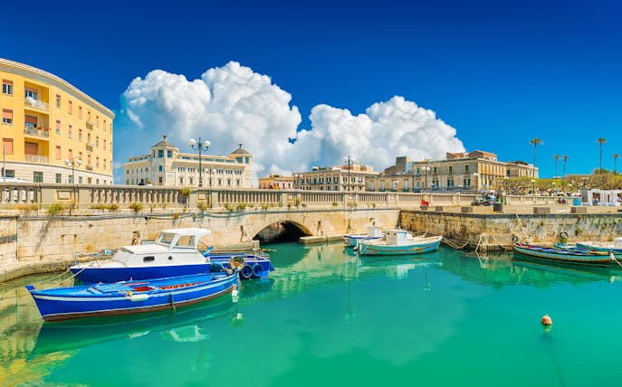 Boats docked in the turquoise waters of Siracusa, Italy, with historic buildings in the background.