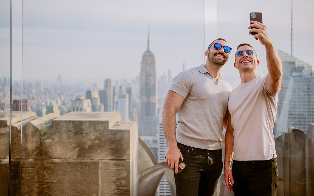 Two people taking a selfie at Top of the Rock Observation Deck with New York City skyline.