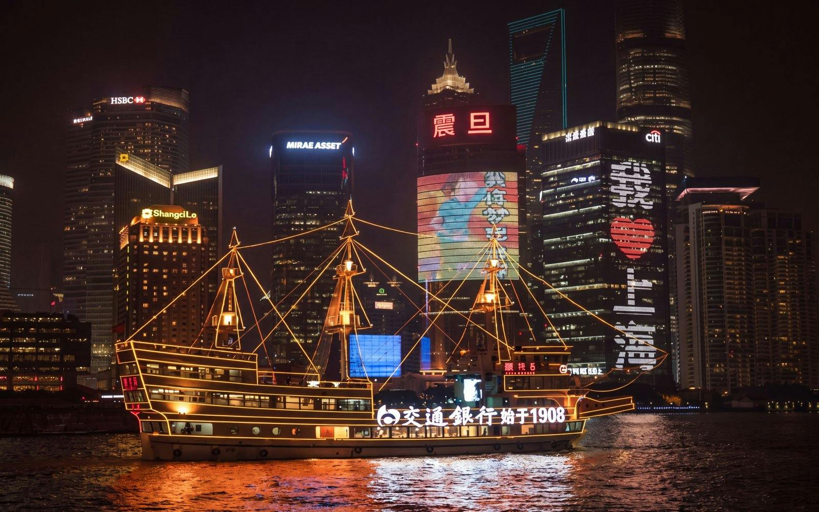 Traditional-style boat illuminated with yellow lights on Huangpu River, Shanghai skyline at night.