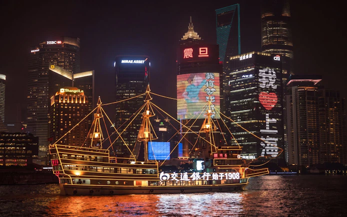 Traditional-style boat illuminated with yellow lights on Huangpu River, Shanghai skyline at night.