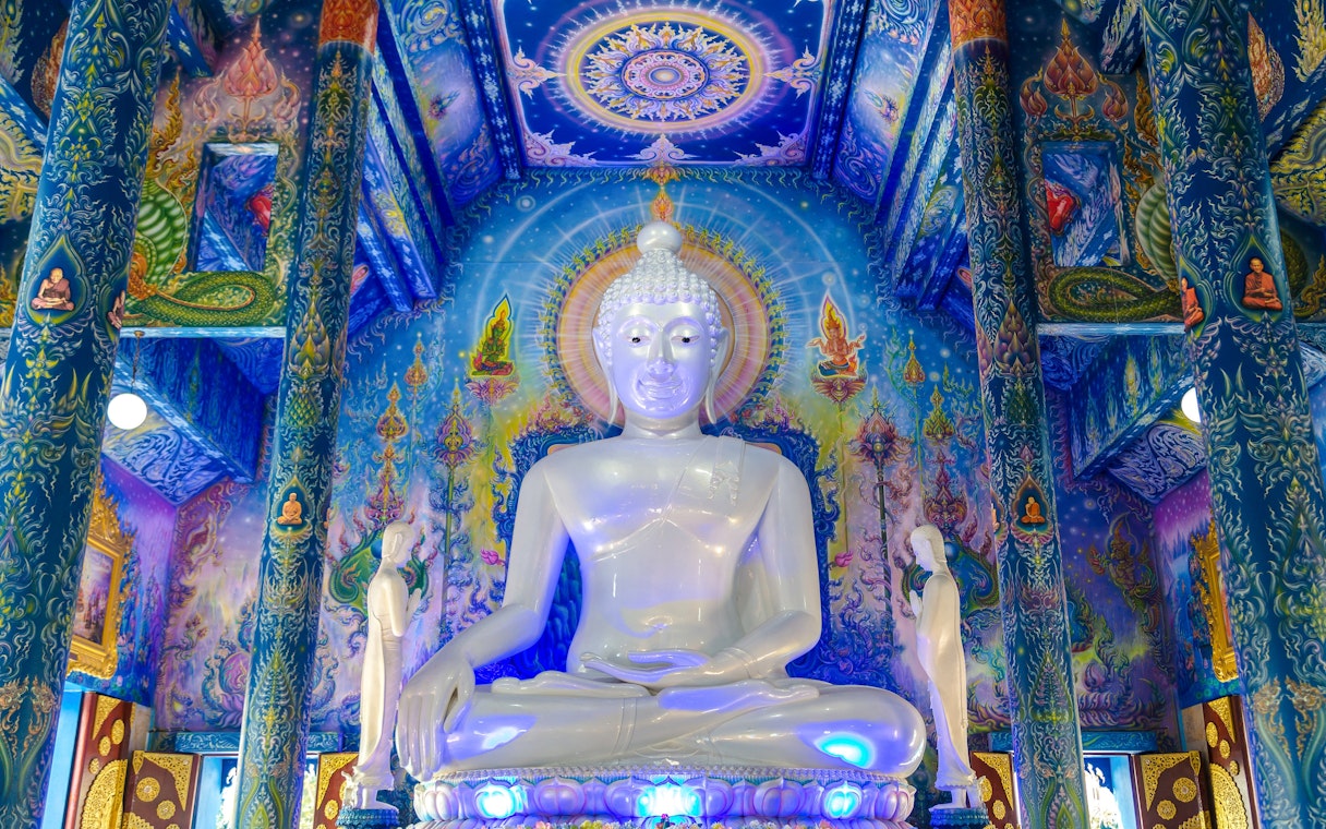 White Buddha statue inside Wat Rong Sua Ten, Chiang Rai, with vibrant blue murals.