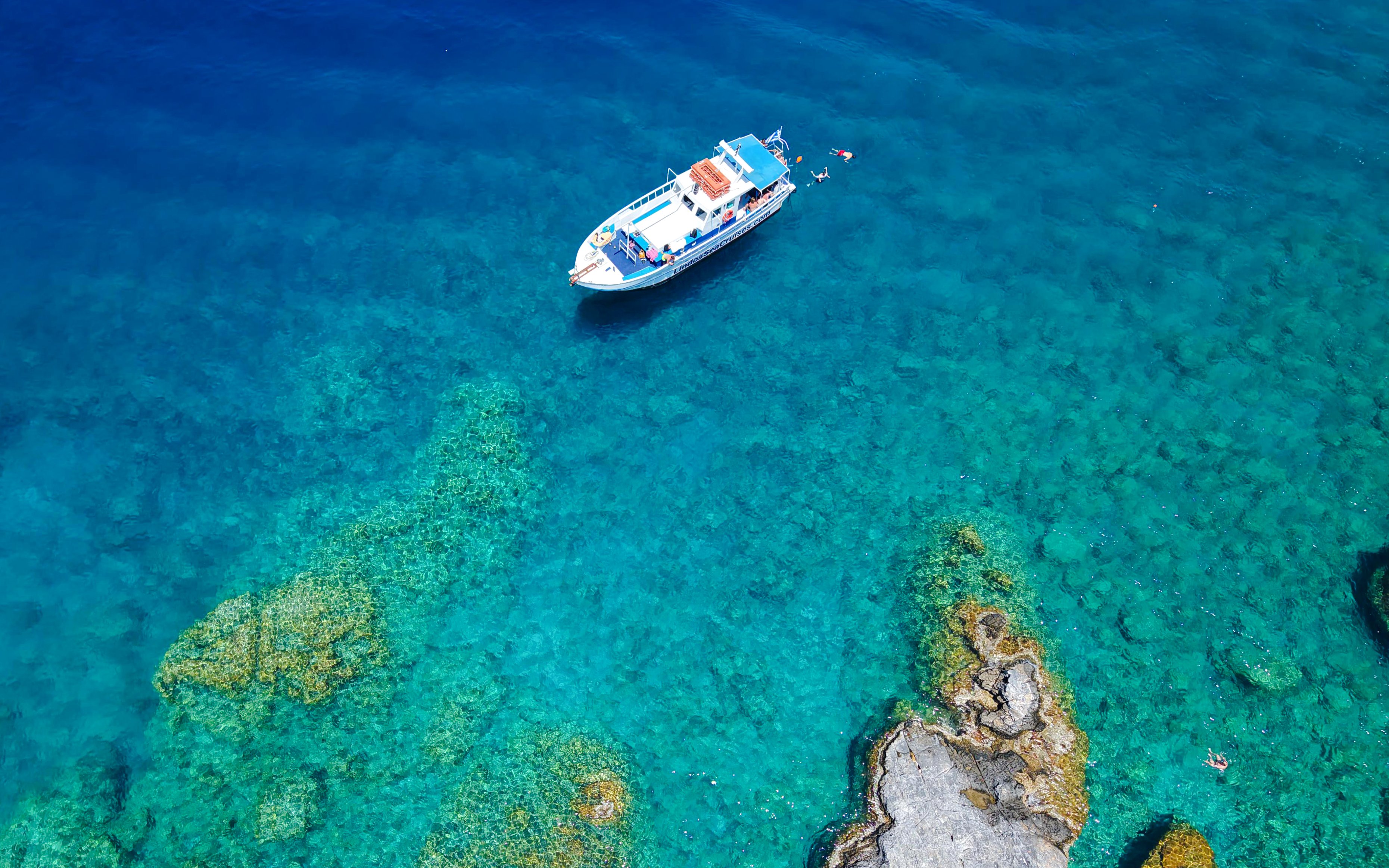 Boat cruising near rocky coastline in Rhodes, Greece, with swimmers in clear blue water.