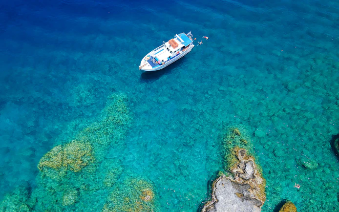 Boat cruising near rocky coastline in Rhodes, Greece, with swimmers in clear blue water.