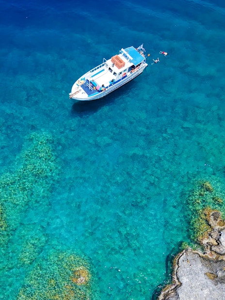 Boat cruising near rocky coastline in Rhodes, Greece, with swimmers in clear blue water.