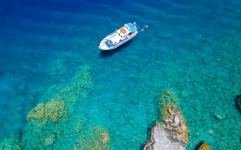 Boat cruising near rocky coastline in Rhodes, Greece, with swimmers in clear blue water.
