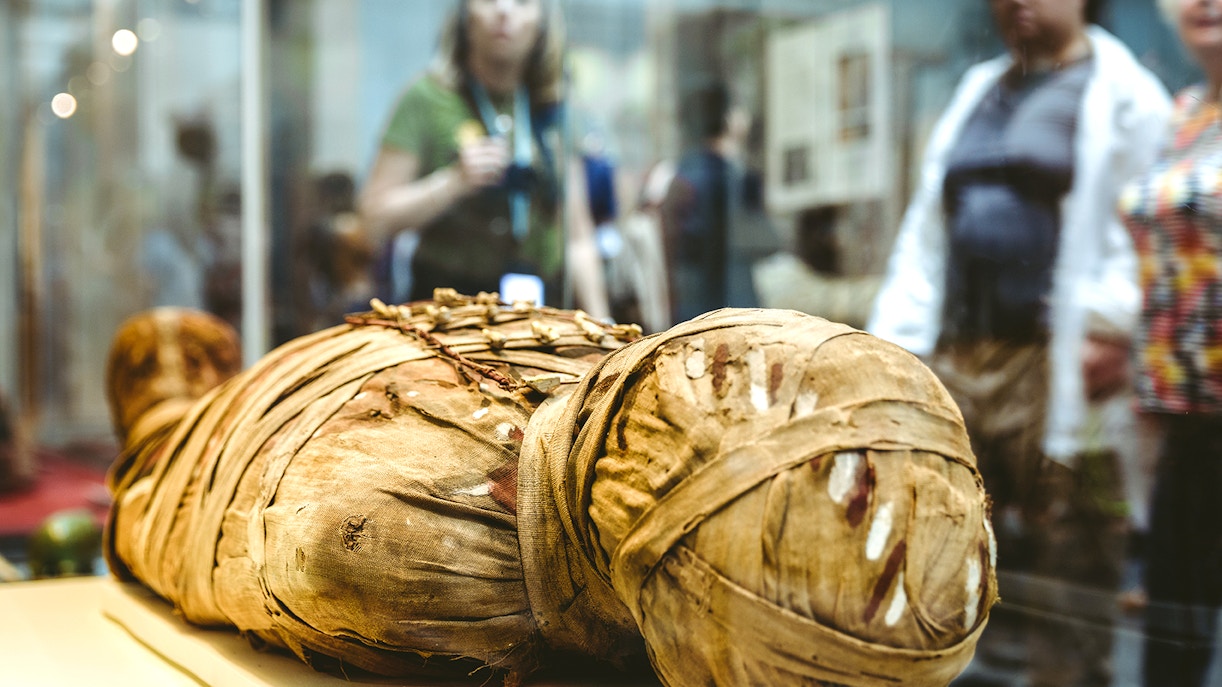 Ancient mummy display at the British Museum with visitors in the background.