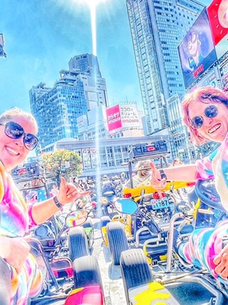 Two women in tie-dye costumes in go-karts at Shibuya Crossing, Tokyo.