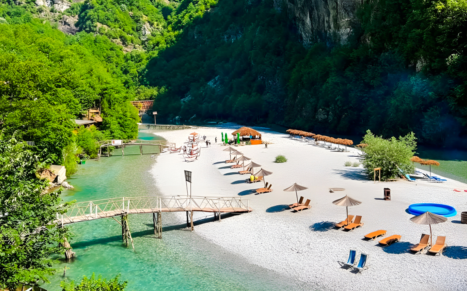 Shala River beach with loungers, umbrellas, and a wooden bridge in a lush green valley.