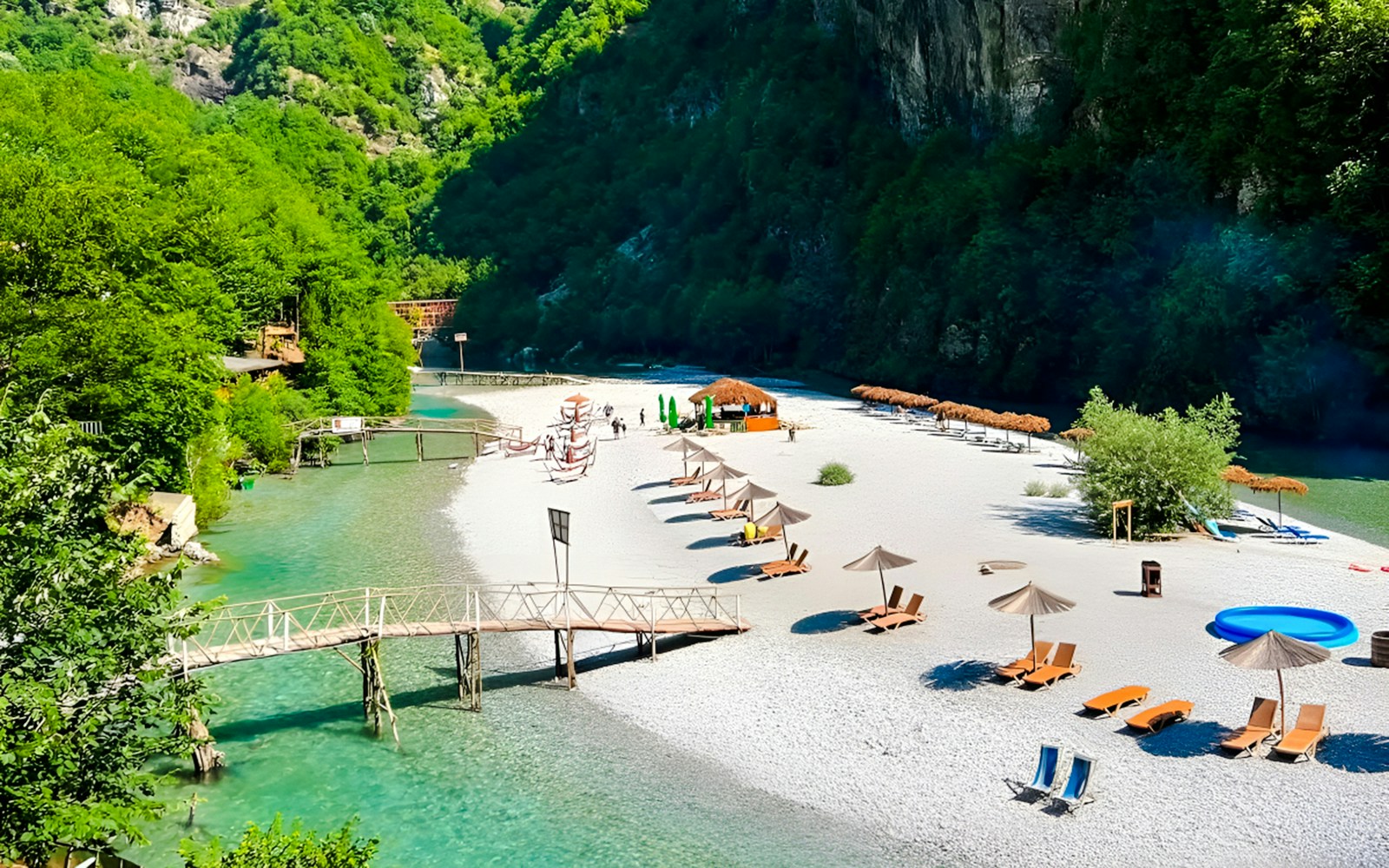 Shala River beach with loungers, umbrellas, and a wooden bridge in a lush green valley.
