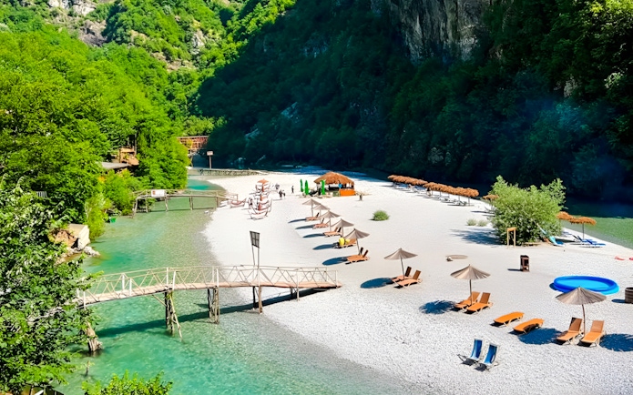 Shala River beach with loungers, umbrellas, and a wooden bridge in a lush green valley.