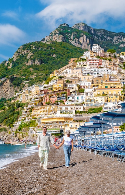 Couple walking on Positano beach with colorful hillside buildings, Amalfi Coast.