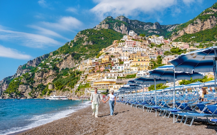 Couple walking on Positano beach with colorful hillside buildings, Amalfi Coast.
