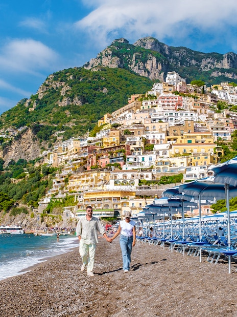Couple walking on Positano beach with colorful hillside buildings, Amalfi Coast.