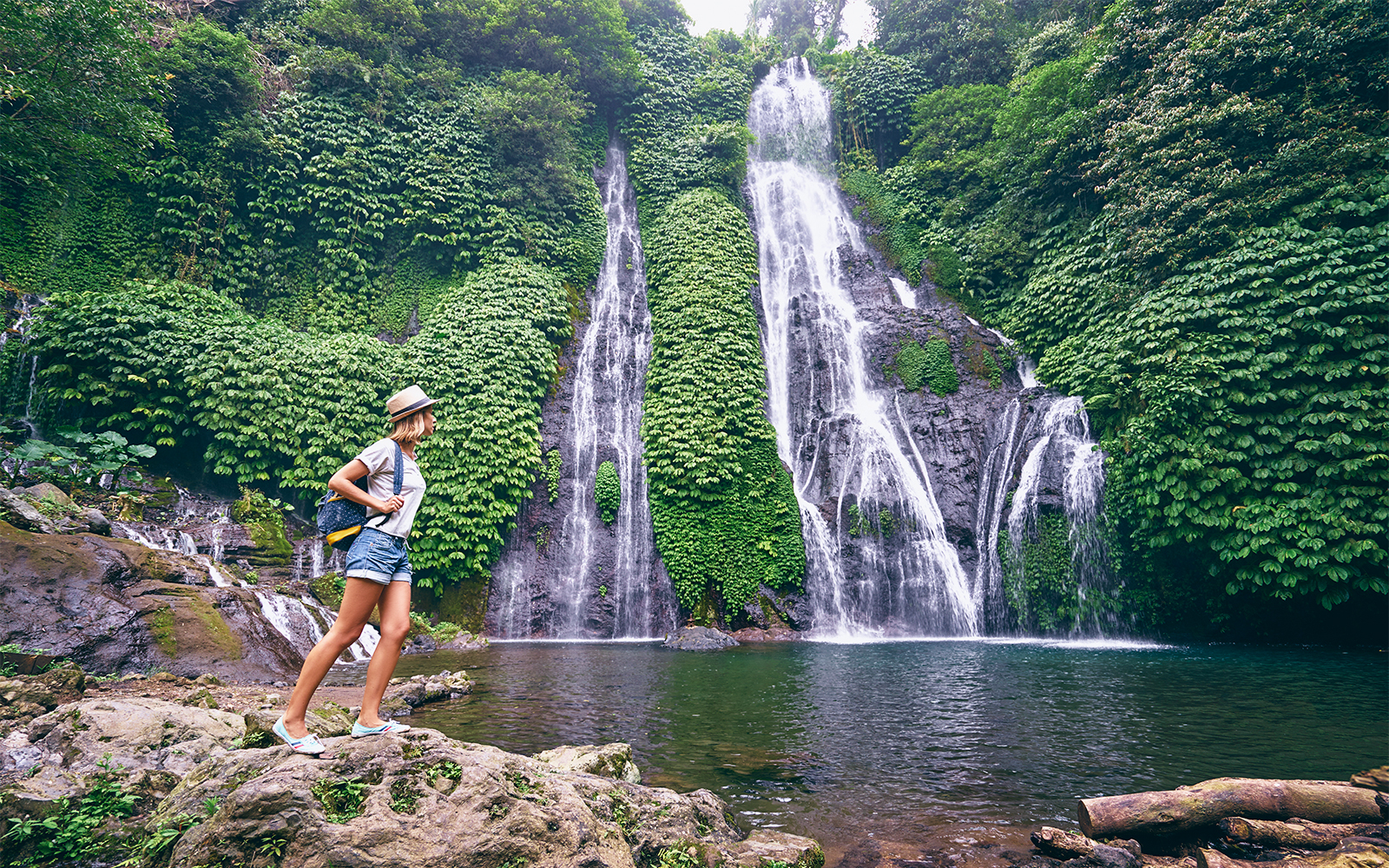 Young woman with a backpack and hat standing near a waterfall in a comfortable dress, exploring nature.