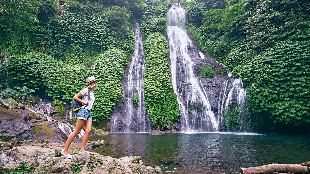 Young woman with a backpack and hat standing near a waterfall in a comfortable dress, exploring nature.