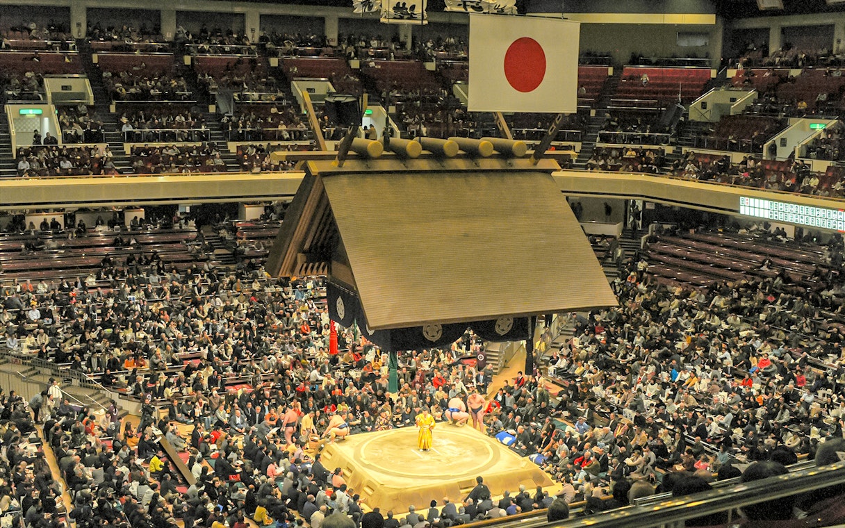 Sumo wrestling match in a crowded Tokyo stadium with a central ring.