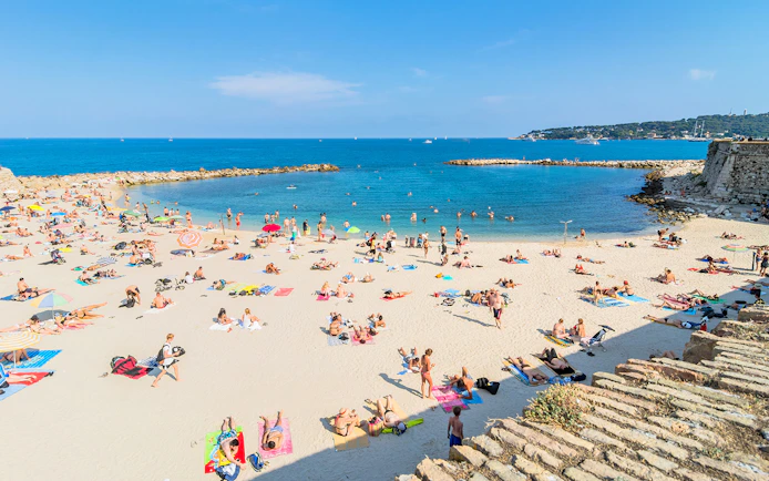 Beachgoers relaxing on a sandy beach in Antibes, France, with clear blue water and distant coastline.